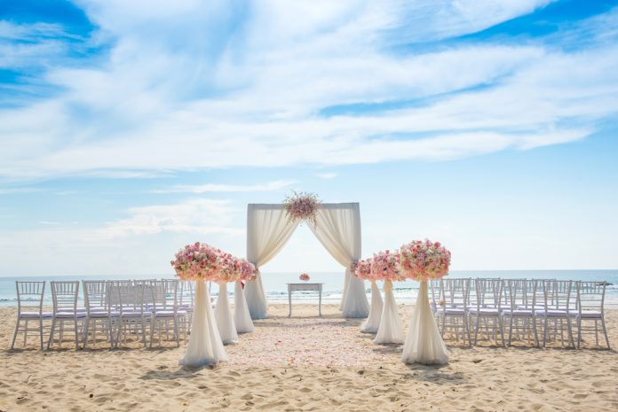 Outdoor beach wedding setup with a white draped arch adorned with pink and white flowers, rows of chairs on either side, and a clear blue sky above.