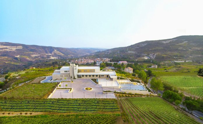 Aerial view of a modern white building surrounded by lush vineyards, with rolling hills in the background under a clear, bright sky.