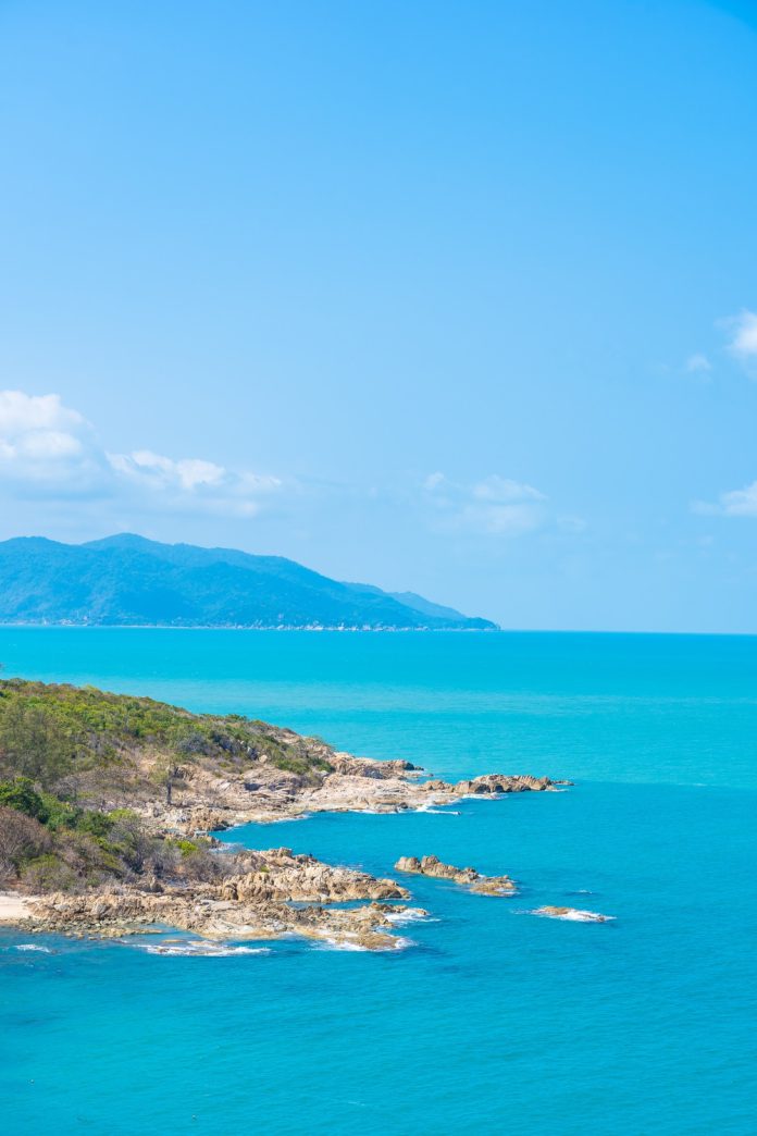 View of a turquoise sea from a rocky coastline with hills and a distant mountain under a clear blue sky.
