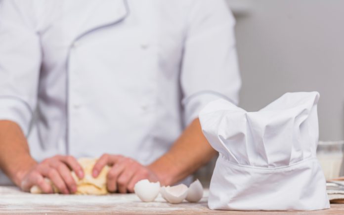 A chef kneading dough on a flour-dusted counter with a white chef's hat and eggshells in the foreground, emphasizing the action and elements of baking.