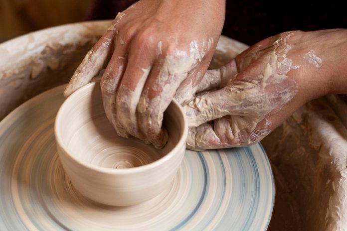 Close-up view of hands shaping clay on a pottery wheel, with the clay spinning into a bowl shape. hands and wheel are covered in wet clay.