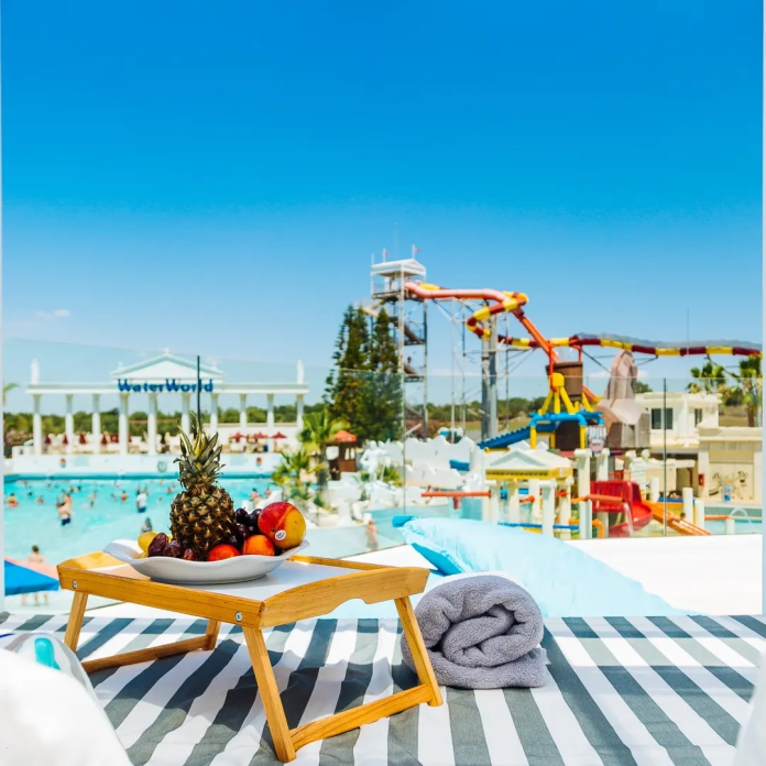 A relaxing poolside scene with a wooden table holding a pineapple and other fruits, and a grey towel on a striped lounge chair, overlooking a vibrant water park with slides.