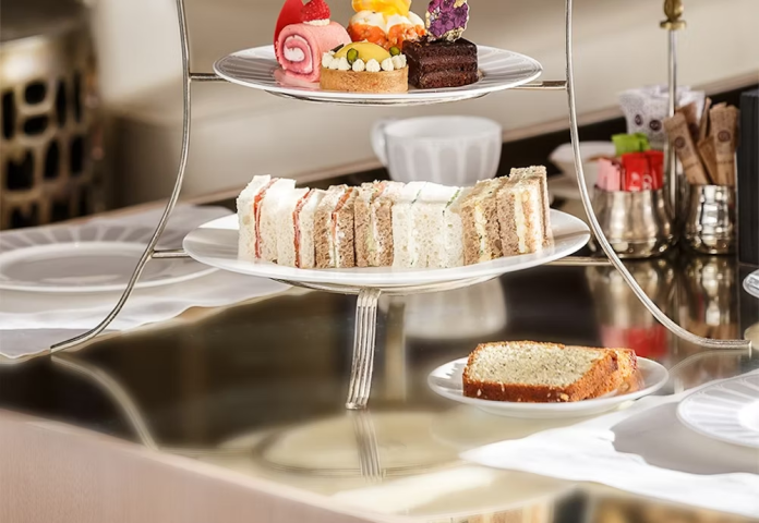 A three-tiered serving tray filled with various types of desserts including slices of cake, colorful frosted cupcakes, and assorted pastries, set on a shiny table in an elegant setting.