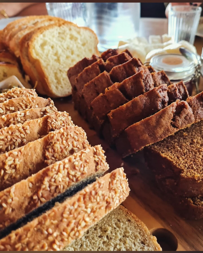 Various types of freshly sliced bread on a wooden table, focusing on one loaf topped with sesame seeds, with other loaves and a small jar of honey in the background.