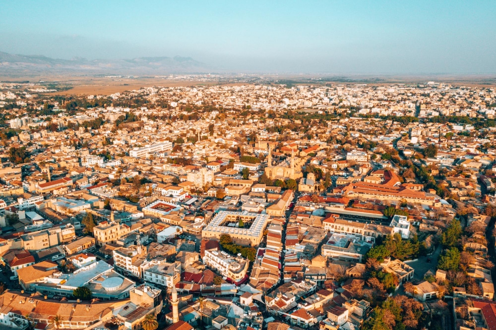 Aerial view of a bustling city at sunset, showcasing densely packed buildings and streets with a prominent historic church at the center surrounded by lush greenery.