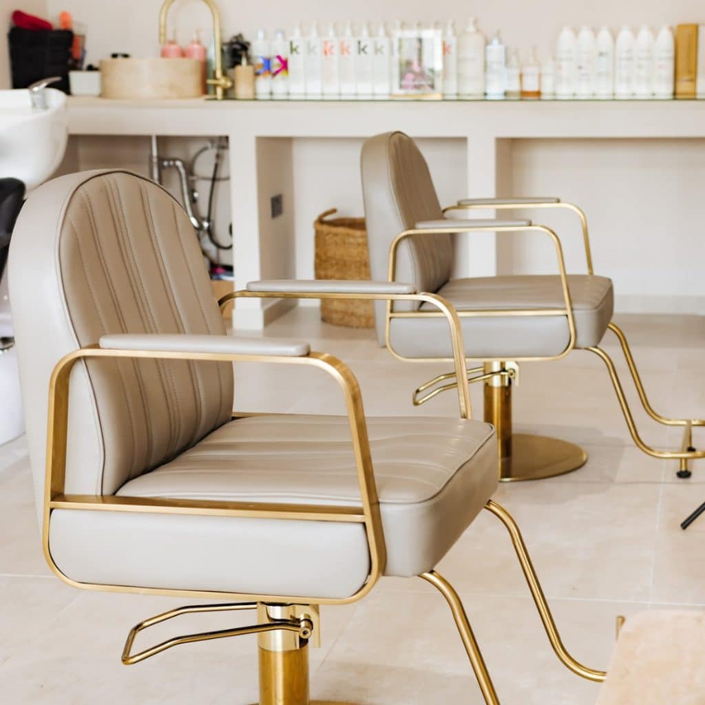 Two stylish salon chairs with beige upholstery and golden frames are arranged on a tiled floor, facing a counter with hair care products on the shelves behind.