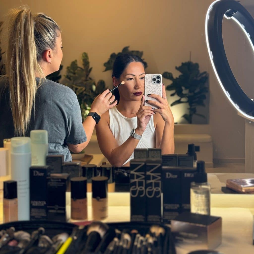 A woman applying makeup in front of a mirror, assisted by another person. products from dior and nars are scattered on the counter. she is holding a smartphone.