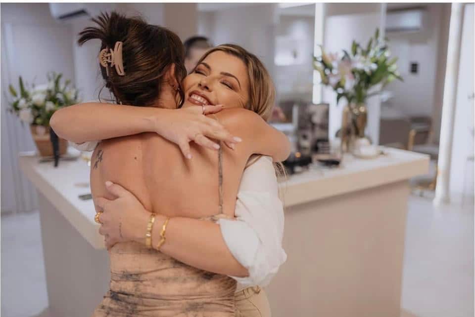 Two women embrace joyfully in a well-lit room with modern decor. one woman, with her back to the camera, has an open-back dress while the other, facing forward, smiles broadly.