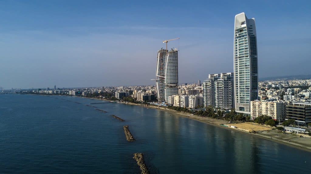 Aerial view of a coastal city skyline featuring modern high-rise buildings, with a clear sky and calm blue sea along a sandy beach.