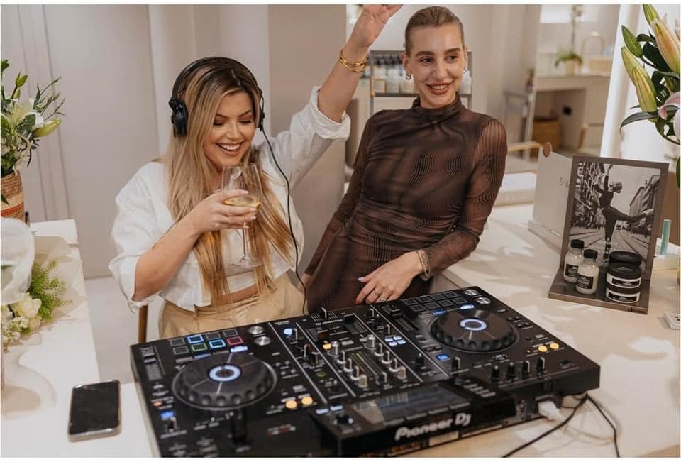 Two women enjoying a moment at a house party, one djing and the other dancing with a drink in hand, surrounded by a festive setup with modern decor.