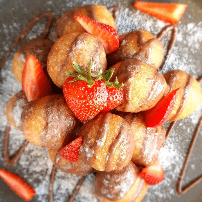 A plate of freshly made mini doughnuts dusted with powdered sugar, drizzled with chocolate sauce, and adorned with sliced fresh strawberries.