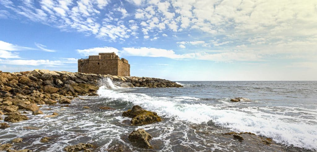 A rugged coastline with rocks and waves crashing ashore, featuring an ancient stone fortress at the edge of the sea under a bright, cloudy sky.