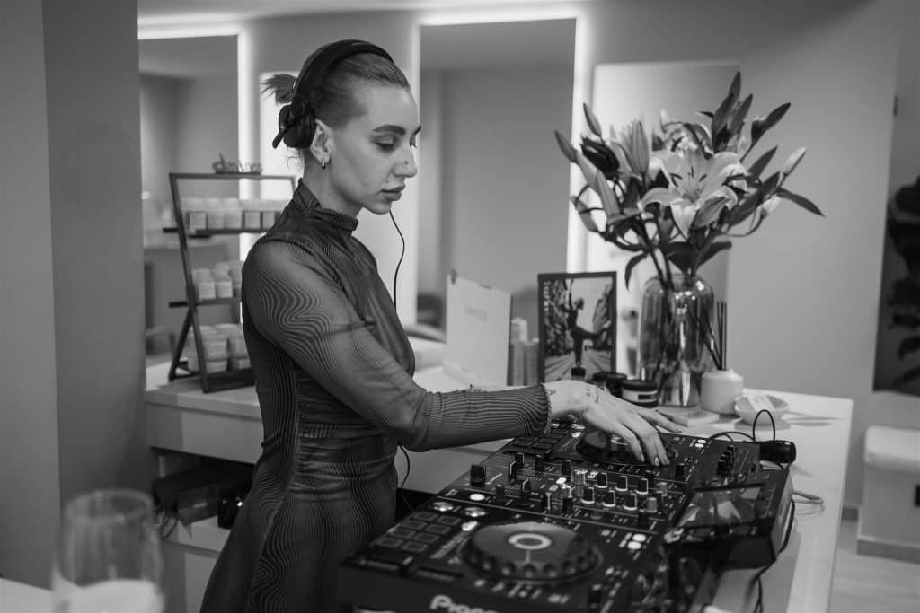 A dj in a sleek, long-sleeve outfit is concentrated on adjusting equipment at her dj setup in a modern room, with flowers and photos in the background. the image is in black and white.