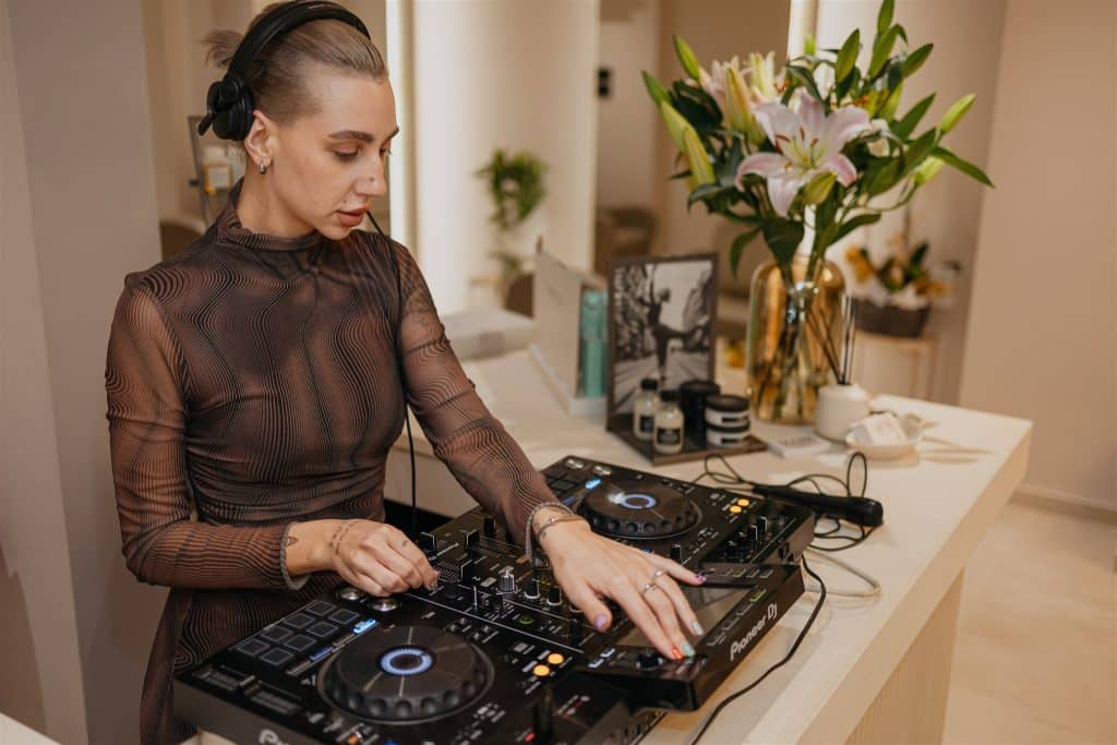 A woman dj with a sleek bun hairstyle mixes tracks on a dj controller at a stylish indoor event space, surrounded by soft lighting and elegant decor.