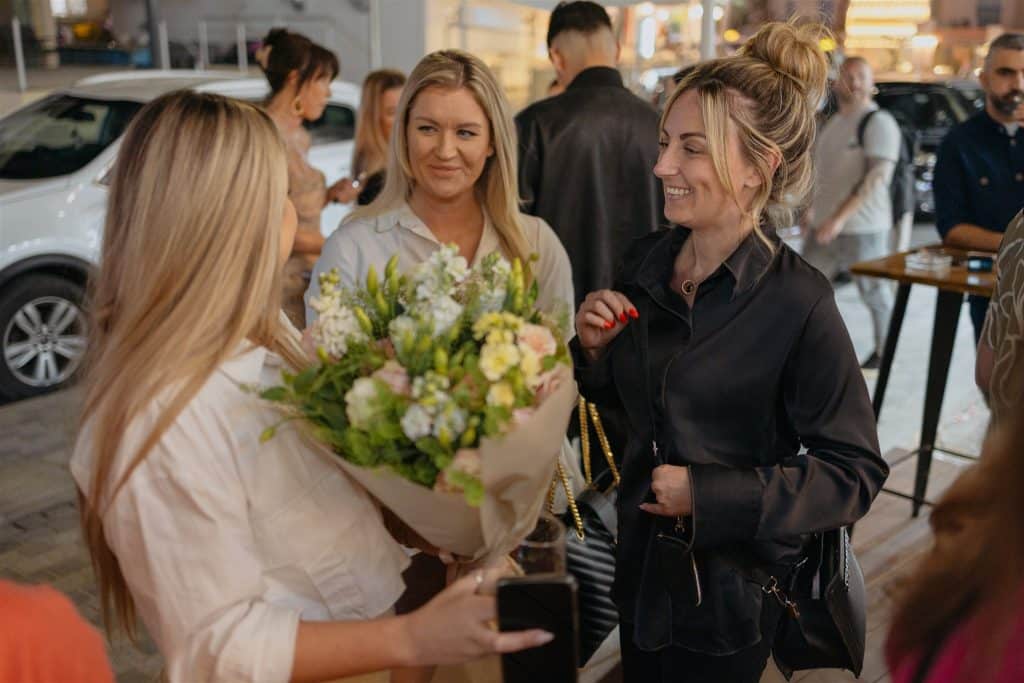 A woman holding a large bouquet of flowers smiles as she greets friends on a lively city street at night, with people casually socializing in the background.