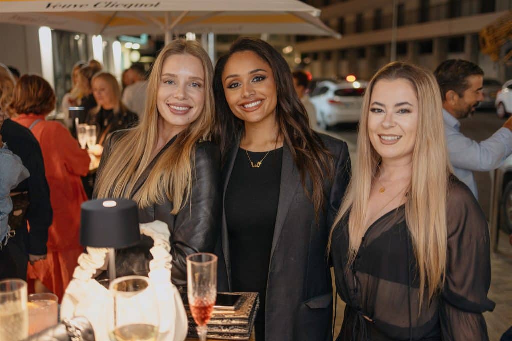 Three women smiling, standing together at an evening social event, dressed in stylish outfits with drinks on a table in front of them.