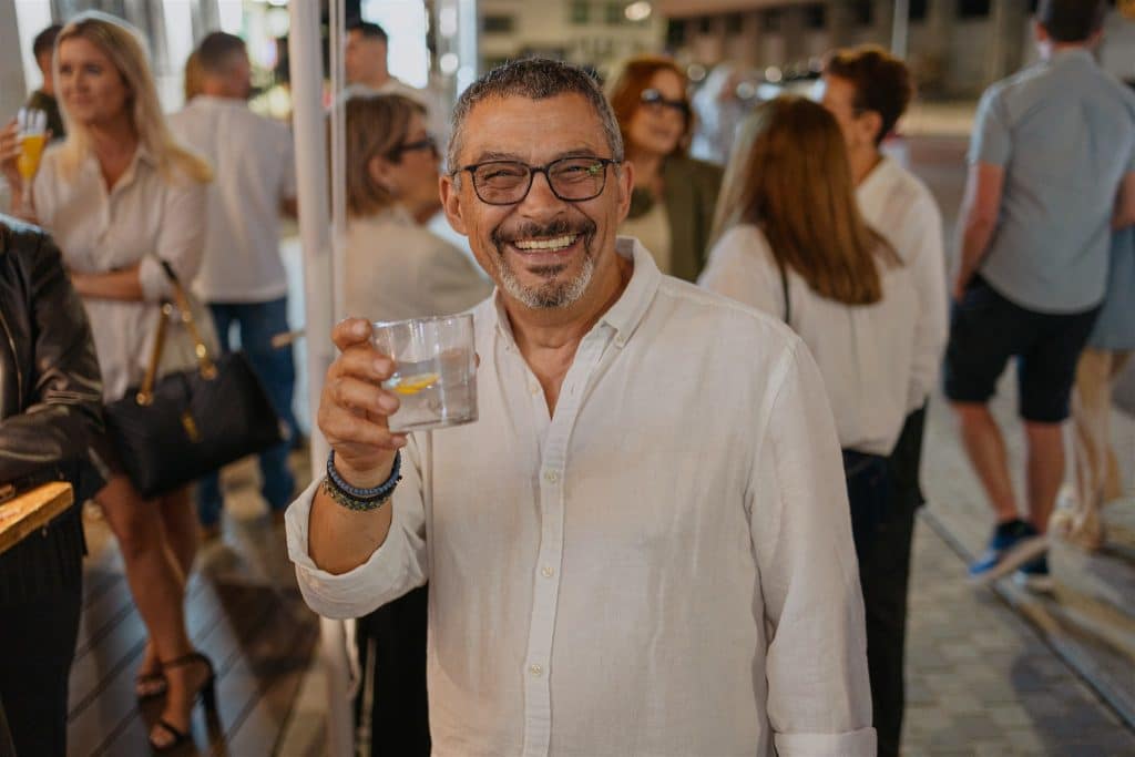 An older man with glasses and a white shirt, smiling joyfully while holding a drink at a street gathering with other people in the background.