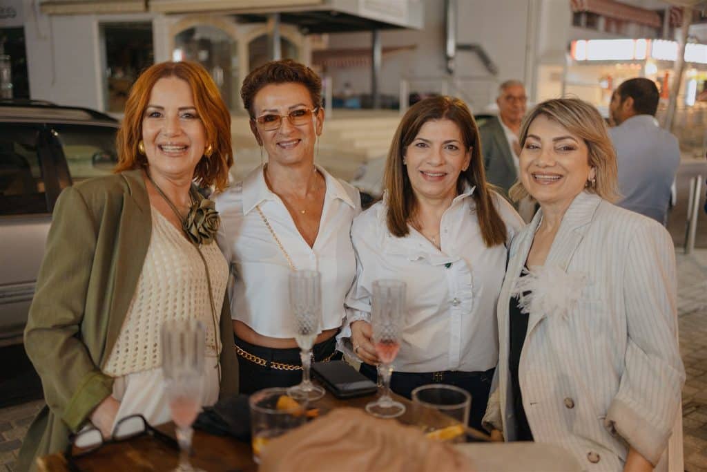 Four women enjoying an evening together, smiling in a social outdoor setting, stylishly dressed, holding drinks and posing for the camera.