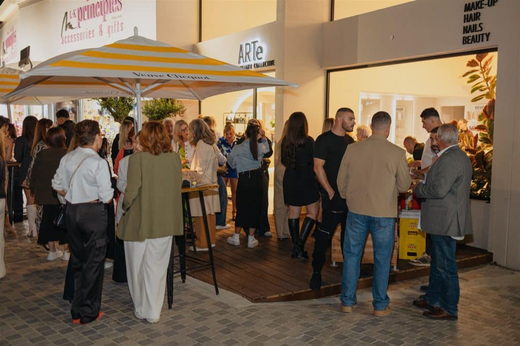 People gathered at a lively outdoor event in front of a shop with indoor lighting visible. attendees chat and enjoy drinks, some standing while others browse inside the shop.