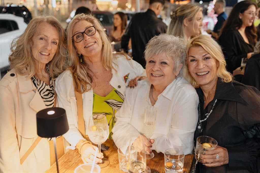 Four senior women smiling and enjoying drinks at a vibrant social gathering, dressed elegantly in a cozy, well-lit setting.