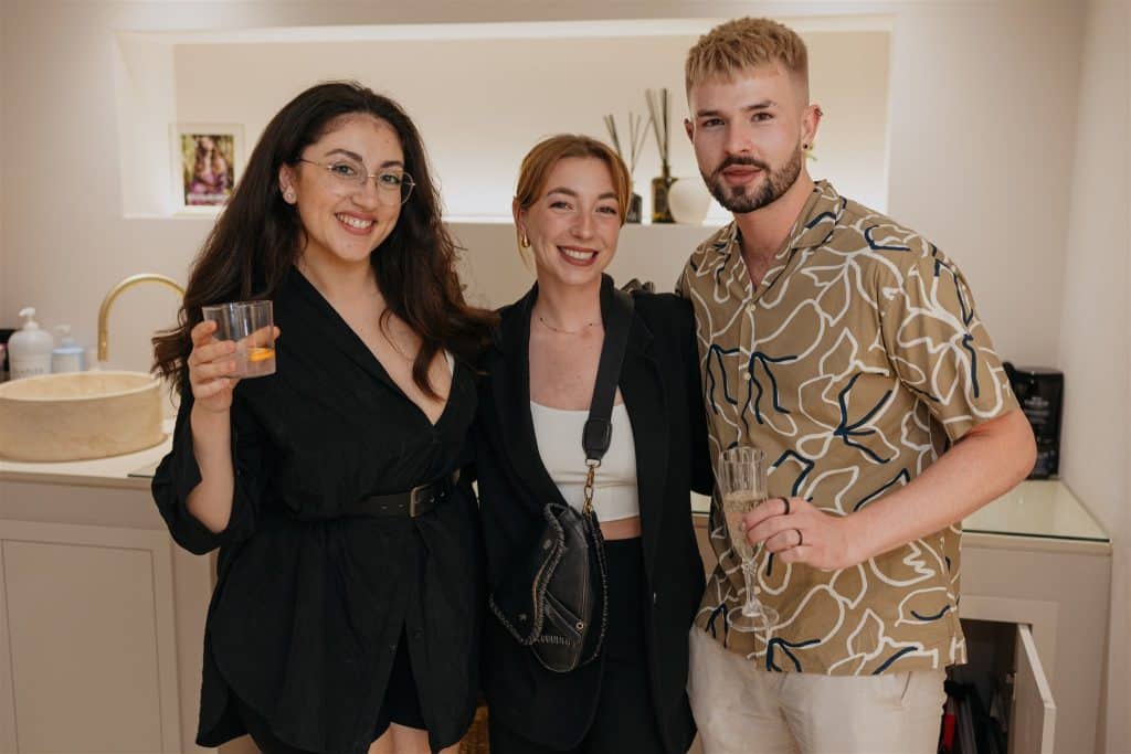 Three young adults, two women and one man, smiling and holding drinks, standing closely together in a warmly lit room with a modern decor.