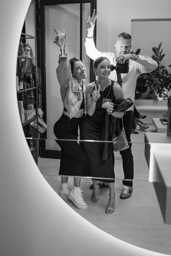 Two women and a man taking a selfie in a stylish interior; one woman raises her hand joyfully while the other points forward, and the man holds the camera.