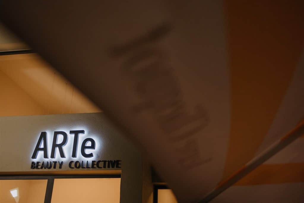 A view of a business sign with the words "arte beauty collective" in backlit white letters, viewed beneath a blurred triangular architectural element at night.