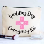 A canvas pouch labeled "wedding day emergency kit" with a pink cross, placed beside makeup essentials including a lipstick, brush, and compact powder on a marble surface.