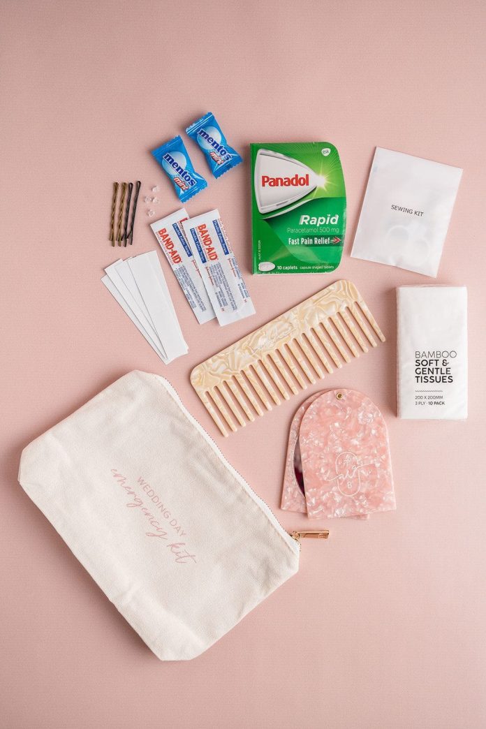 Emergency kit with items like pain relief tablets, bandages, sewing kit, bamboo tissues, soap, and a comb spread out beside a canvas bag on a pink background.